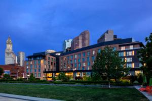 Modern hotel at dusk with downtown Boston buildings behind it and Rose Kennedy Greenway Park in front.
