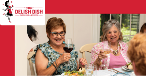 Two happy women enjoying themselves at a holiday event with a stylized logo for the upscale Cincinnati area catering company, The Delish Dish, to the left of them.
