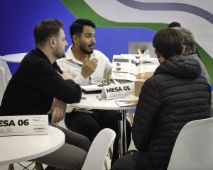 Four professionals are gathered around a table during a business networking session in a trade fair environment. Two men are facing the camera, while two participants have their backs turned. One of the professionals, wearing a white shirt, is gesturing w