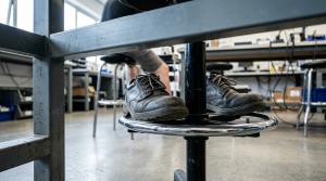 Work shoes resting on the chrome adjustable footring of a draughtsman chair at an ESD electronics bench, with the workshop floor clearly out of reach below