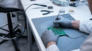 A technician soldering a circuit board at a raised ESD workbench, with a draughtsman chair and its adjustable footring visible at the edge of the frame