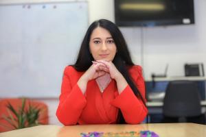 Tammy Kassiou with long dark hair, wearing a red top, resting her chin on her hands and smiling at a desk in an office setting.