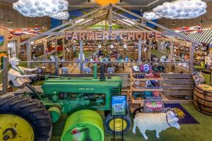 A timber barn frame under a lit Farmers Choice marquee, with a vintage tractor, a mannequin driver, a sheep figure and hanging cloud lights.
