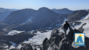 Person standing on a rocky mountain ridge overlooking a rugged, snow-covered alpine landscape, with the GoWild Wilderness Adventures logo in the lower-right corner.