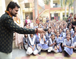 Adv. Peeyush Pandit, wearing a dark patterned jacket and a marigold garland, speaks to a large group of smiling schoolgirls in blue-and-white uniforms gathered around him in a school courtyard.