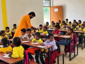 Adv. Peeyush Pandit, wearing an orange kurta, speaks with young students seated at desks with notebooks inside a classroom in India.