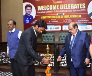 Adv. Peeyush Pandit, dressed in a dark suit, bends forward to light a traditional brass ceremonial lamp at an International Conference on Early Childhood Care & Education at the Constitution Club of India in New Delhi. Two men stand nearby as he lights th