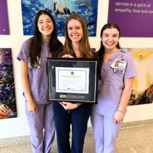 Three smiling women in purple scrubs holding a framed Certified Autism Center® award in a colorful art-decorated room.