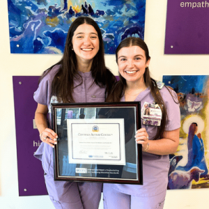 Two women in purple scrubs smiling and holding a framed "Certified Autism Center" certificate in front of colorful artwork.