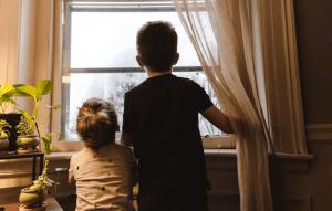 Two young children standing near a window with sheer curtains.