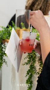 A guest holds a pink beverage in an It’s A Secret Med Spa cup with a syringe-shaped stirrer and a branded gift bag.