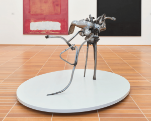 A steel abstract sculpture balancing on three spindled legs within the center of a museum gallery with colorful paintings in the background