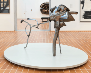 A steel abstract sculpture balancing on three spindled legs within the center of a museum gallery with colorful paintings in the background