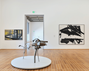A steel abstract sculpture balancing on three spindled legs within the center of a museum gallery with colorful paintings in the background