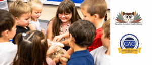 A group of children happily gathered around a woman holding a stuffed animal in a classroom setting.