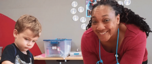 A woman smiling at the camera while a young boy next to her focuses on an activity at a classroom table.