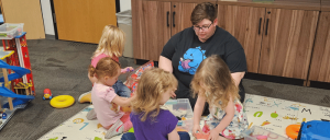 Adult reading a book to four young children sitting in a playroom on a colorful alphabet mat with toys around.