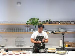 Wenwen co-founder and creative chef Eric Sze demonstrates a cooking technique behind a kitchen counter during a culinary presentation.