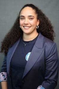 Shanie Harvie, EDP, Director of Client Success and Implementation at Economic Impact Catalyst, smiling in a professional headshot. She has long, dark curly hair and wears a navy blazer with a floral-lined cuff over a navy top, against a gray studio backdr