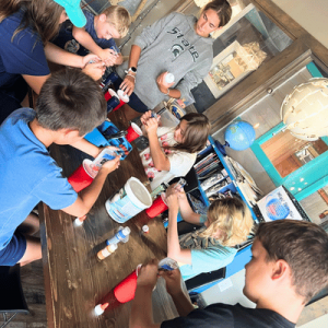 A group of children gathered around a table painting and decorating red plastic cups in a classroom setting.