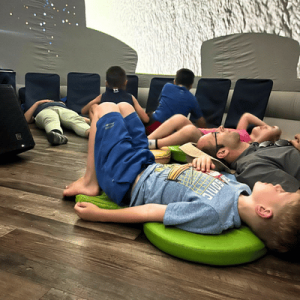 Children lying on cushions on a wooden floor inside a dome-shaped room with cushioned chairs against the wall.