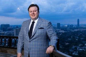 Alexander Vitenas standing on a Houston balcony in a gray plaid suit with the city skyline behind him.