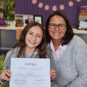Young girl holding a LearningRx certificate, smiling next to a woman in glasses, both seated in a cozy, decorated room.