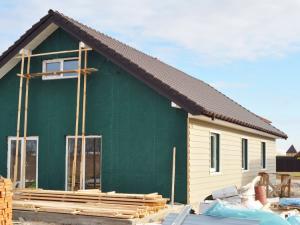 Partially sided house showing cream lap siding on the side wall and green house wrap on the front gable, with wood scaffolding, lumber and building materials on the ground.