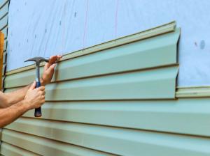 Close up of a worker's hands holding a hammer while fitting a pale green vinyl siding panel onto a wall, with exposed sheathing and chalk layout lines visible above the finished courses.