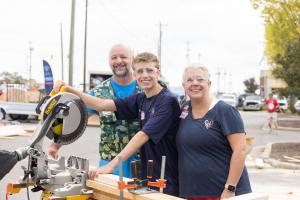 A Lowe's associate helps a mother and her teenage son operate a miter saw at an outdoor bed-building event.