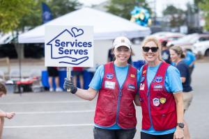 Two Lowe's associates in red service vests smile together, one holding a "Services Serves" sign, in a store parking lot set up for a bed-building event.