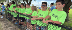 A group of children in bright green T-shirts stand along a black chain-link fence outdoors under trees on a sunny day.