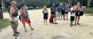A group of people wearing life jackets stand on a gravel path near parked cars, preparing for an outdoor water activity.
