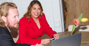 The image shows the two founders of Dandelion, a legacy and estate planning platform. Amanda Brown, a woman in a red suit, and Sam Dalton, a man in a navy jacket, are pictured speaking and smiling across a table.