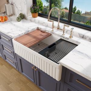 White fireclay farmhouse sink with a vertically fluted apron front set into deep blue cabinets under marble, a wooden cutting board and black drying rack on the ledge, gold bridge faucet above.