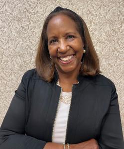 A black woman wearing a black suit and white shirt has her arms folded and is smiling.