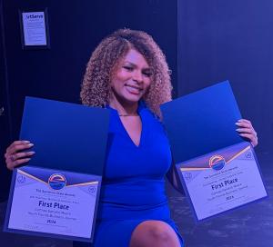 Journalist Cortney Danielle Moore, donning a blue dress, holds up her two first-place certificates at Sunshine State Awards for work she published in 2025. One certificate is for Business and Consumer Affairs while the other is for Lifestyle, Trends & Cul