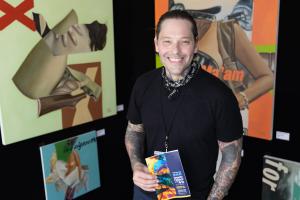 Shows a smiling artist in his booth with his work in the background