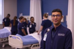 Galen student standing in the foreground while other students learn at the bedside in the background.
