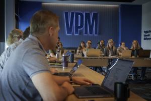 A group of people sitting around tables with open laptops during a meeting in a conference room featuring a prominent illuminated "VPM" sign on the back wall.