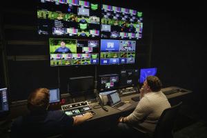 Two production technicians sitting at control boards in a dark control room, facing a wall of multi-view video monitors displaying various camera feeds and a VPM graphic.