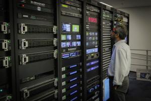 A man looking up at tall server and equipment racks filled with broadcast technology hardware, video monitors, and a digital clock displaying 9:58:05.
