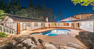Nighttime exterior of a Mediterranean-style estate with a curved pool and spa in a landscaped courtyard, surrounded by mature trees and tile-roofed wings of the home.