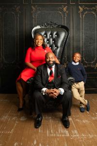 Warm family portrait of a smiling Black mother in red, father in a black suit and red tie, and young son posing by an ornate leather chair.