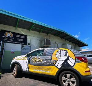 Alpha Omega Plumbing branded car wrap listing drain descaling, drain cleaning, backflow testing, and septic pumping services, parked outside the shop.