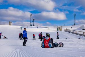 Young snowboarders and an instructor on a groomed ski run on a sunny day, with several youth in red SOS Outreach bibs preparing to snowboard.
