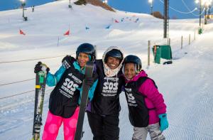 Three smiling skiers in SOS Outreach vests — an adult instructor and two children — pose together on a ski slope at Brighton Resort at dusk, with ski lift towers and slalom gates visible in the background.
