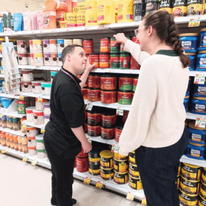 Two shoppers examine cans of paint on a hardware store shelf.