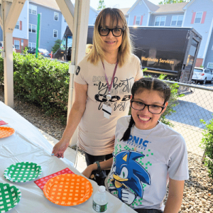 A woman and a smiling girl pose beside a table displaying colorful woven plates outdoors.