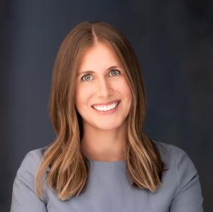 Professional corporate headshot of Cassie Katz standing against a dark studio background.